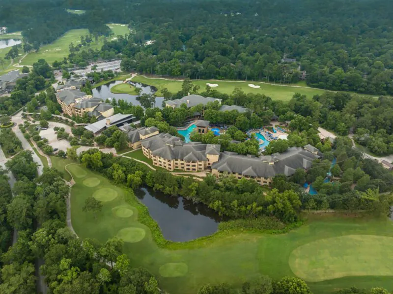 Aerial view of a large resort surrounded by trees, golf courses, pools, and a pond, with winding paths and roads visible.