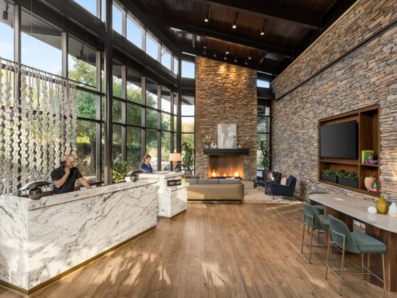 Modern hotel lobby with stone walls, marble desks, a fireplace, large windows, and seating areas. Two people work at reception.