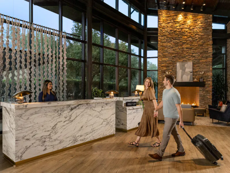 A couple holding hands walks through a hotel lobby; the man pulls a suitcase, and a fireplace glows in the background.