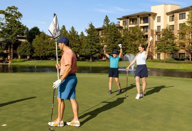 Three people play golf near a pond; one holds a flag while two celebrate with raised arms. Buildings and trees are in the background.
