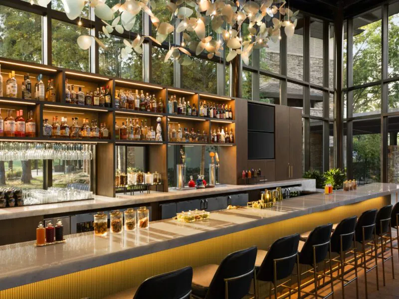 Modern bar with a long counter, black chairs, shelves full of bottles, large windows, and unique ceiling lights. Trees visible outside.