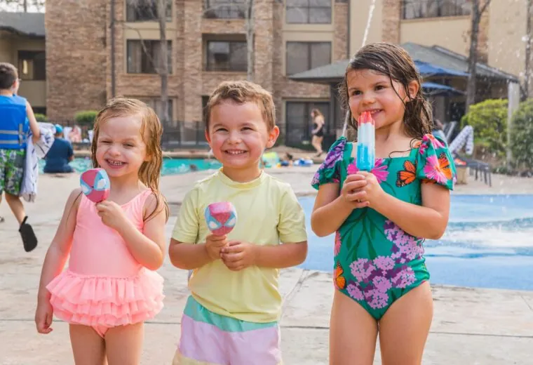 Three young children in swimsuits smile and hold popsicles by a pool on a sunny day, with water features and a building in the background.
