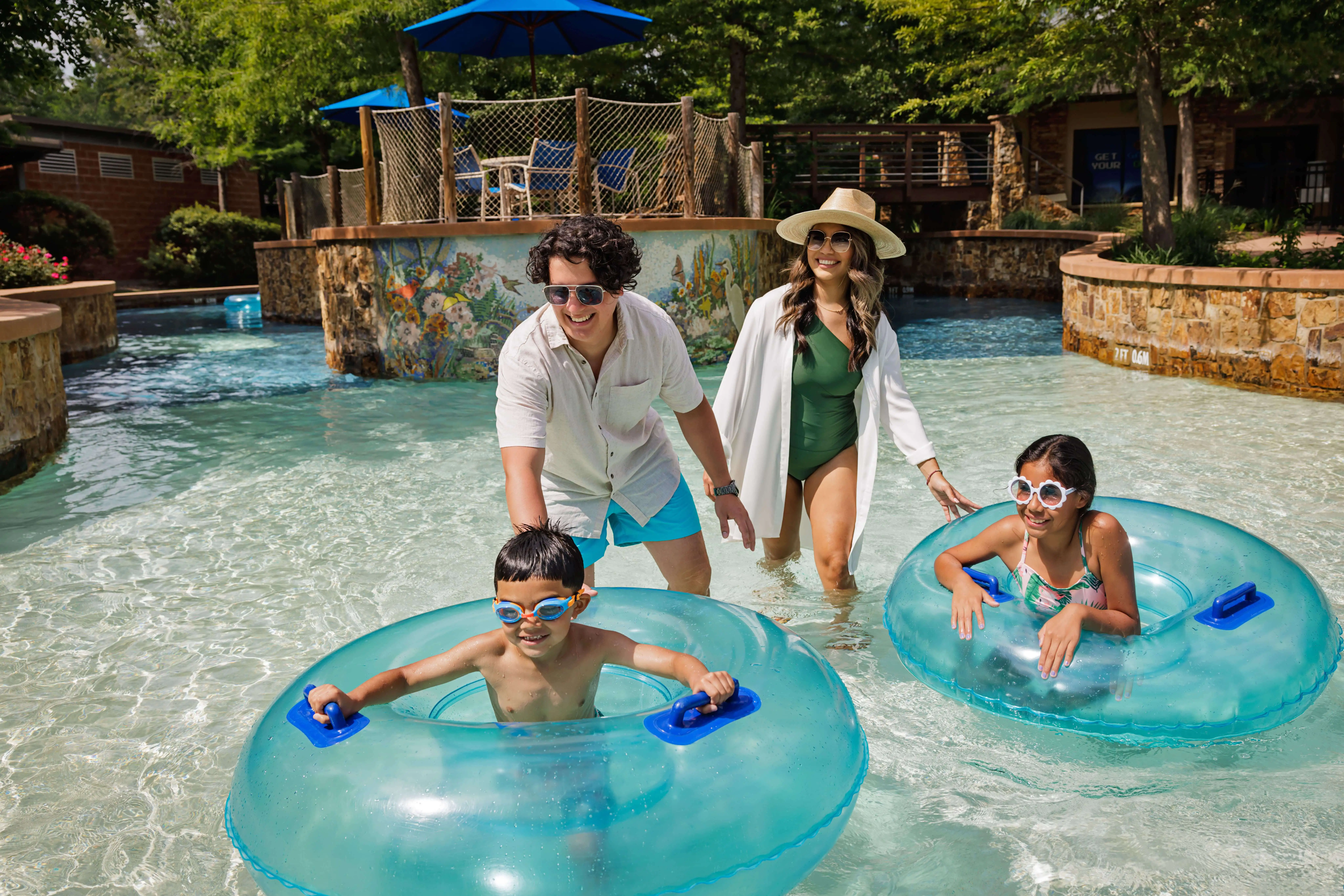 A family of four enjoys a lazy river, with two kids in blue tubes and two adults walking in shallow water, all smiling and wearing swimsuits.