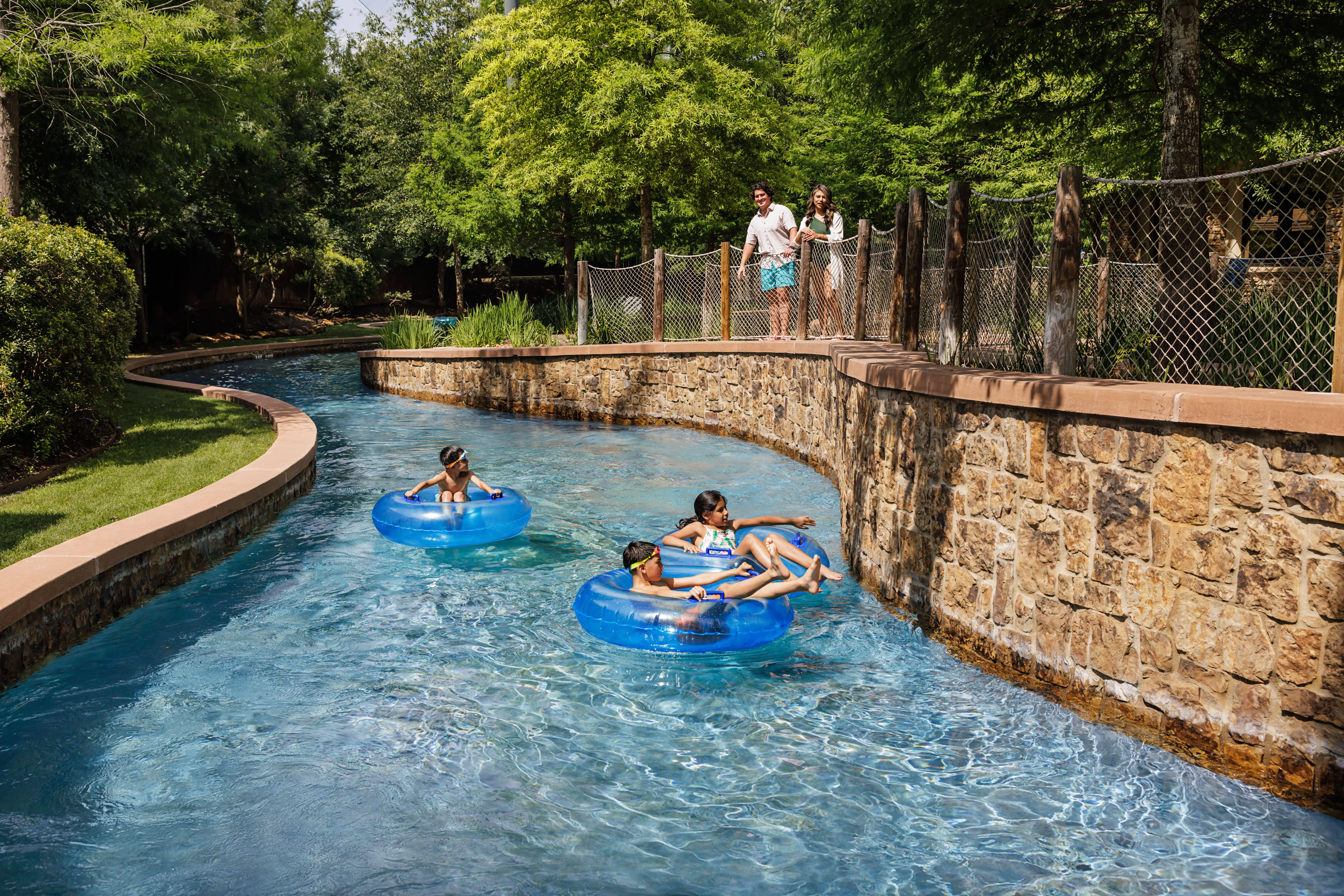 Four children float in blue inner tubes on a lazy river, while two adults watch from a path above, surrounded by greenery and trees.