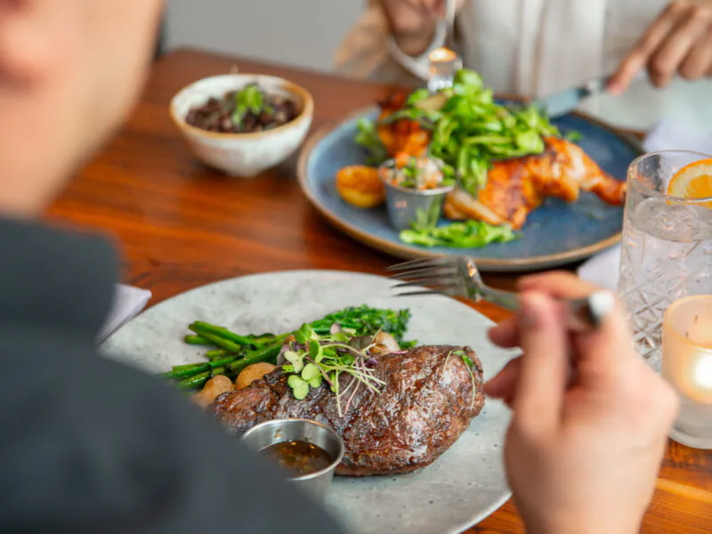 Person dining at a restaurant with a steak, roasted vegetables, sauce, and a drink on the table. Another person eats chicken in the background.