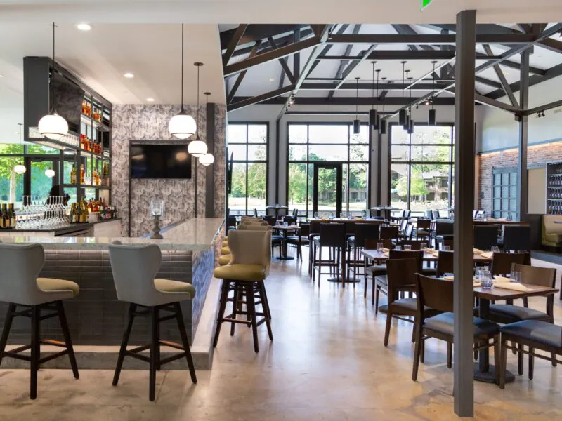 Modern restaurant with a marble bar, high chairs, tables, and large windows letting in natural light. Contemporary decor with exposed beams.