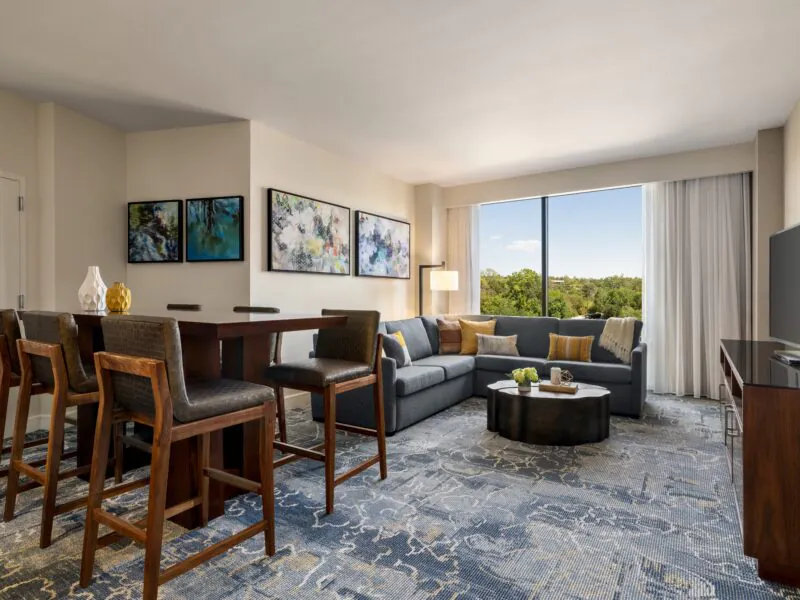Modern hotel suite with dining area, gray sofa, coffee table, large window with a view, and wall art above the couch.