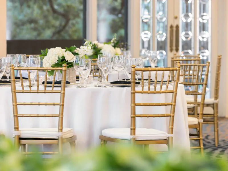 A round table with white linens, gold chairs, glassware, and white floral centerpieces set for an elegant event in a bright room.