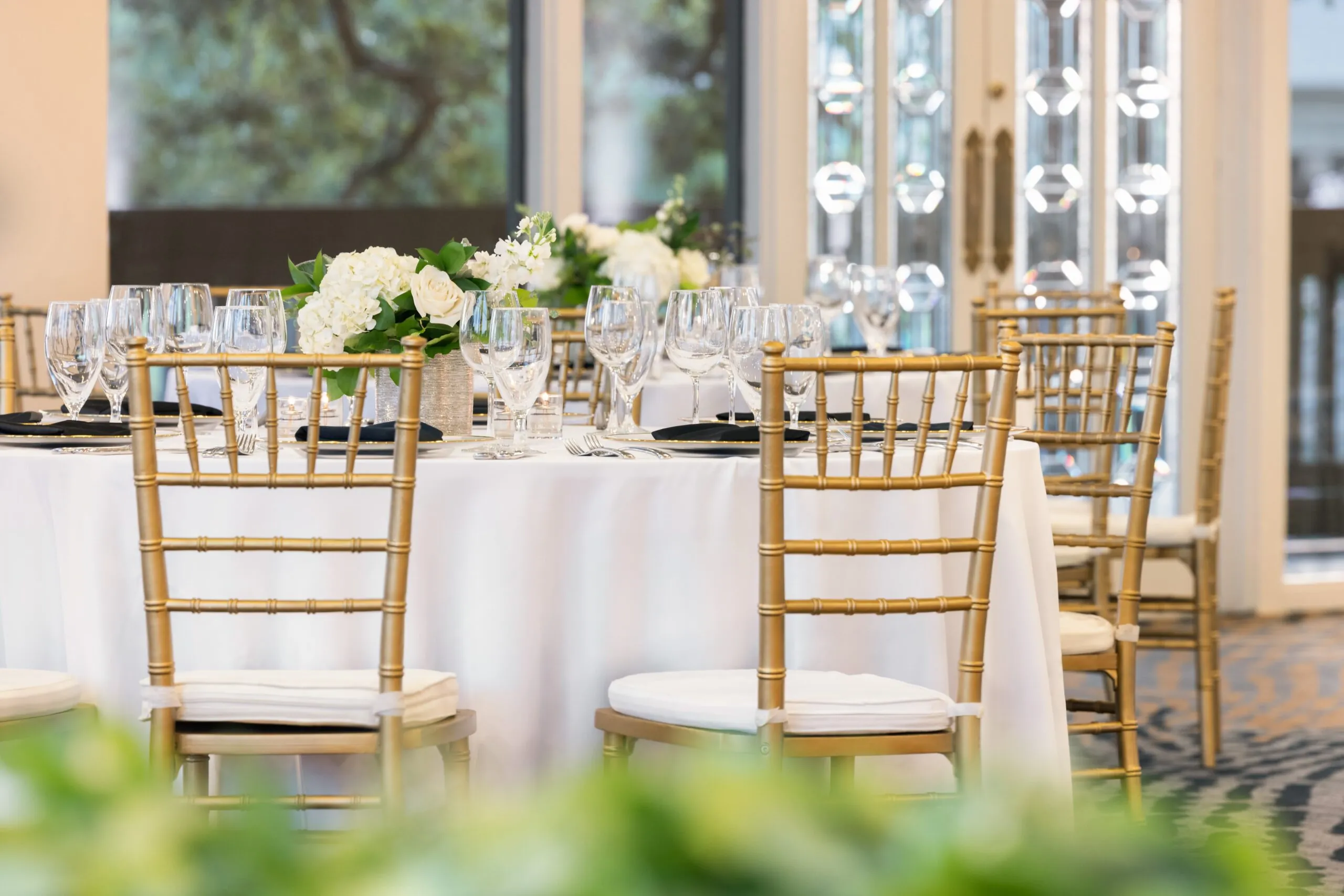 A round table with white linens, gold chairs, glassware, and white floral centerpieces set for an elegant event in a bright room.
