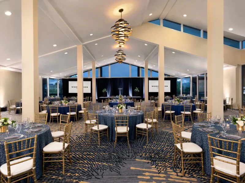 Elegant banquet hall with blue tablecloths, gold chairs, modern chandeliers, and large windows, set up for a formal event or wedding reception.