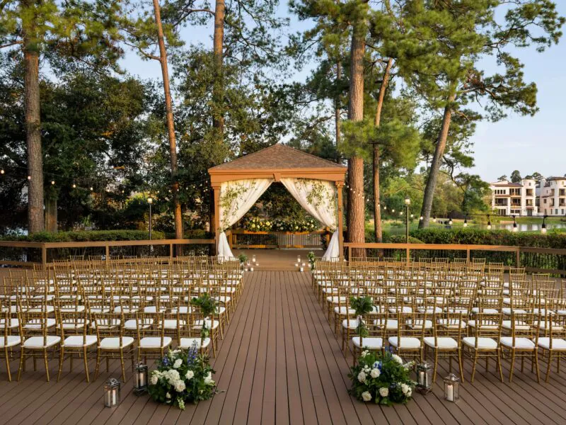 Rows of gold chairs face a draped gazebo on a wooden deck, surrounded by trees and floral arrangements, set for an outdoor ceremony.