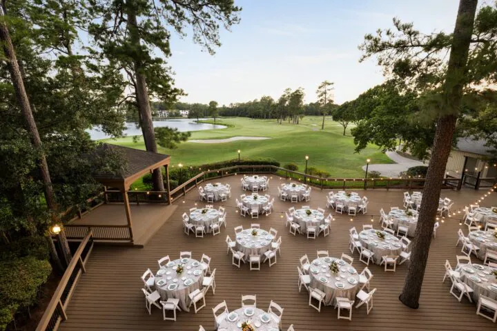 Round tables with white chairs set for an outdoor event on a wooden deck overlook a golf course with trees and a pond in the background.