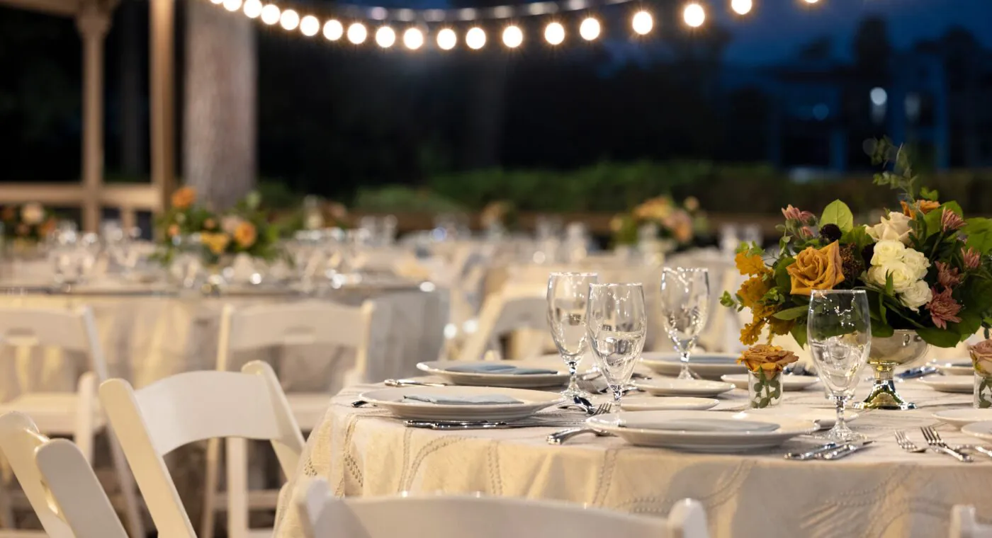 Elegant outdoor event setup with round tables, white chairs, floral centerpieces, and string lights glowing overhead in the evening.