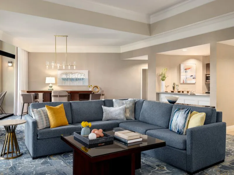 Modern living room with a gray sectional sofa, yellow and white pillows, coffee table, dining area, and open kitchen in a neutral color palette.