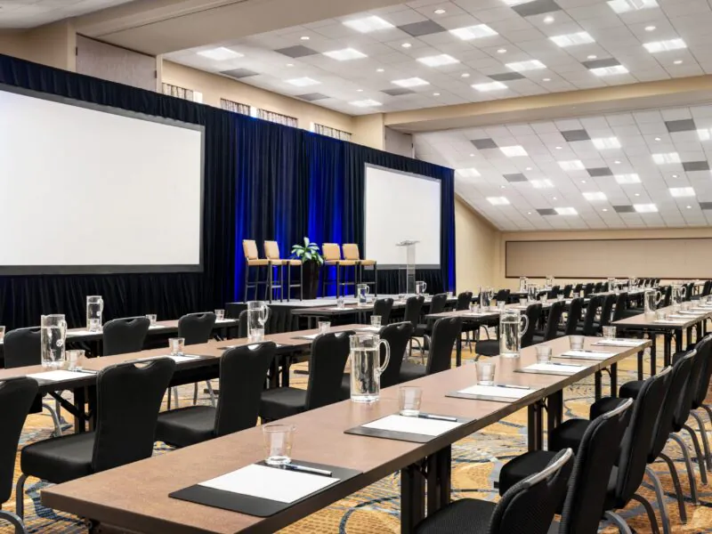 Rows of tables and chairs face two large projection screens and a stage with chairs and a podium in a well-lit conference room.