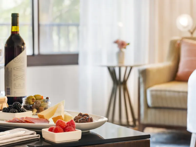 A plate of cheese, fruit, and nuts with a bottle of red wine on a table in a bright, cozy room with a chair and window in the background.
