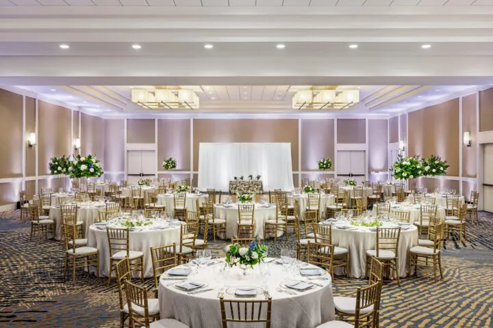Elegant banquet hall with round tables, gold chairs, white linens, floral centerpieces, and a white backdrop, ready for a formal event or wedding.