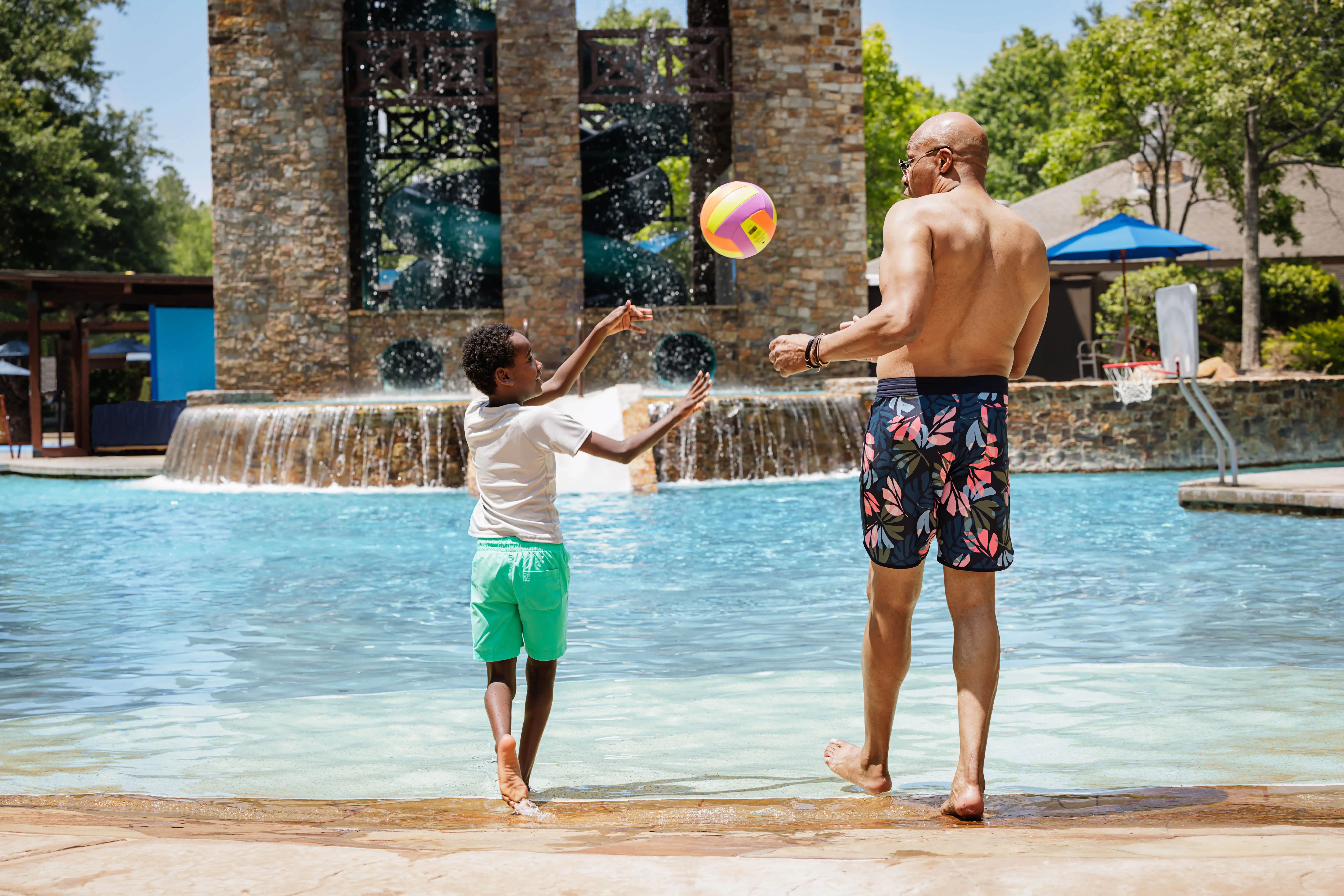 A man and a boy play with a beach ball at the edge of a pool, with fountains and trees in the background on a sunny day.