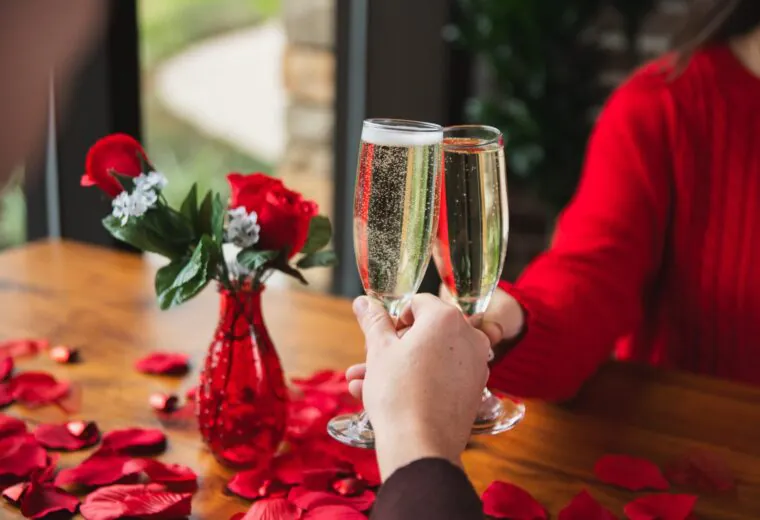 Two people clink champagne glasses over a table with red rose petals and a vase of red roses, creating a romantic atmosphere.