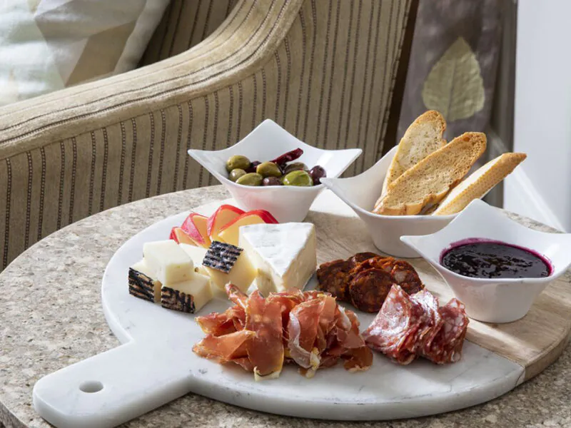 A charcuterie board with cheeses, cured meats, crackers, olives, and berry sauce on a round marble platter next to a beige striped chair.