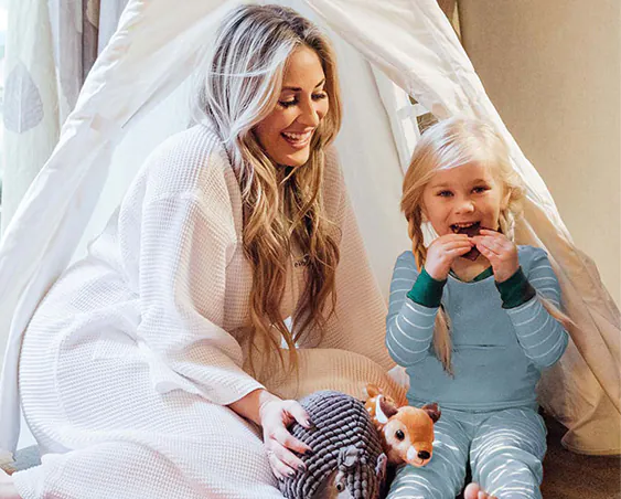 Smiling woman in a white robe sits next to a young girl in blue pajamas eating a snack inside a cozy tent with stuffed animals.