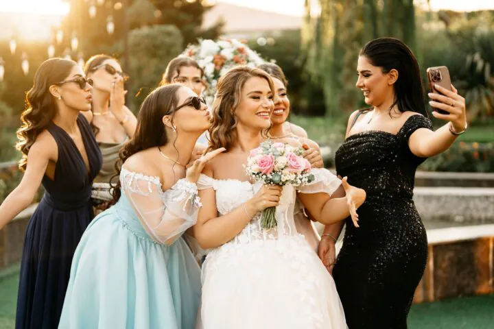 A bride in a white dress holds a bouquet and smiles as her bridesmaids pose and take a selfie with her outdoors.