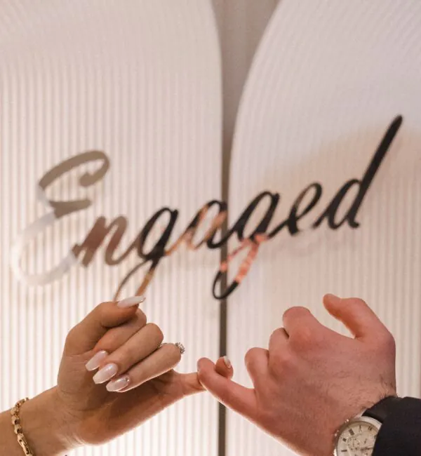 Two hands touch pinkies in front of a sign that says Engaged. One hand has an engagement ring, the other wears a watch.