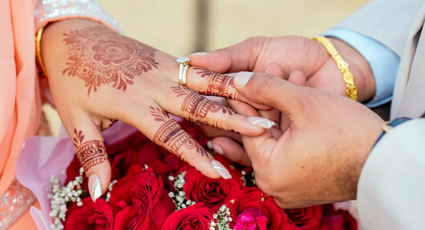 A person places a ring on anothers finger, both hands have henna designs. A bouquet of red roses is in the foreground.