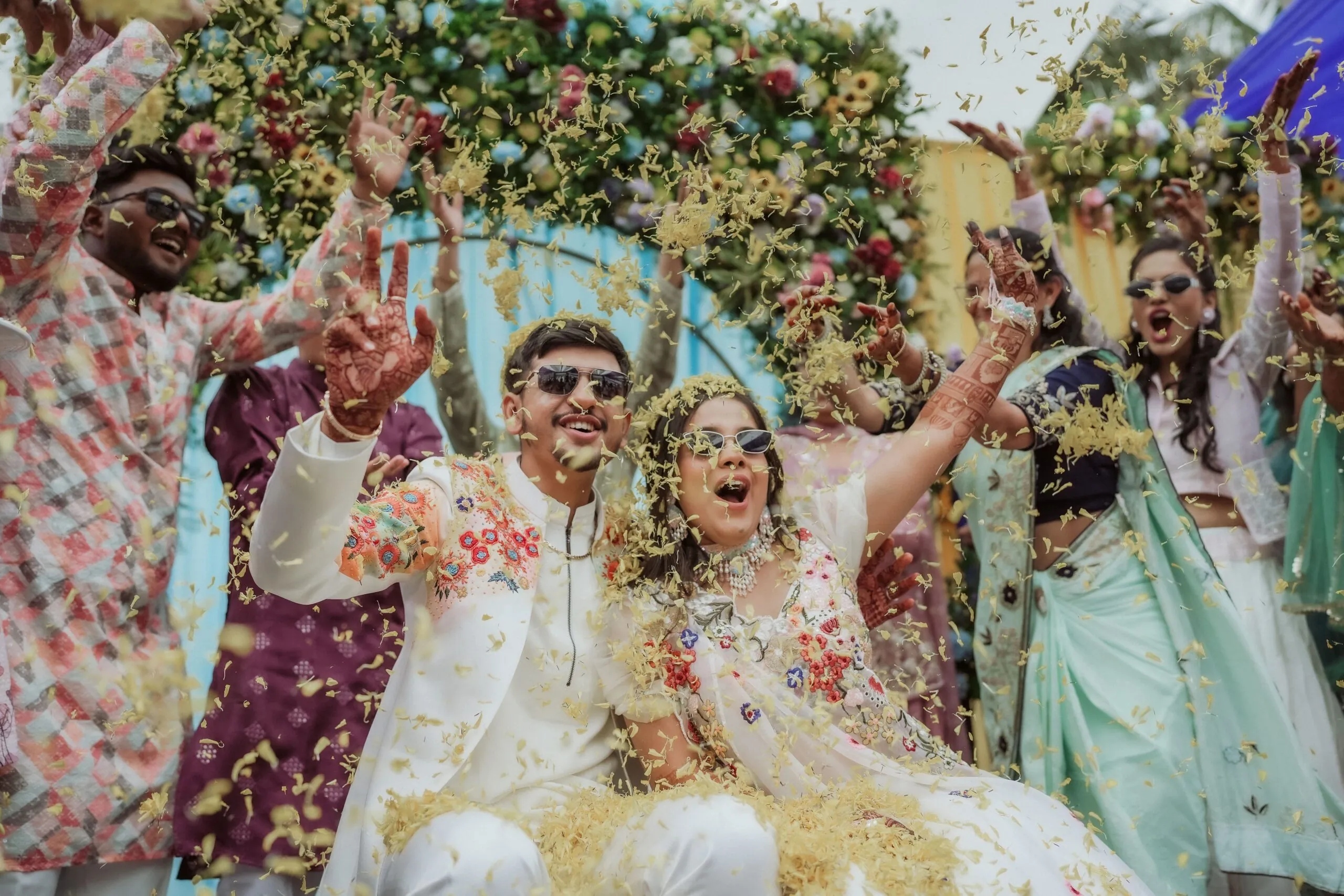 A joyful couple in traditional attire celebrate with friends, tossing flower petals in the air at a colorful outdoor event.