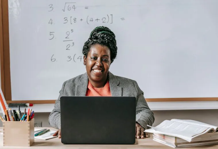 Smiling person sits at a desk with a laptop and open book, in front of a whiteboard with math problems written on it.