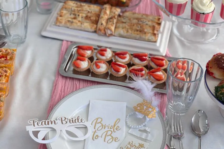 A bridal shower table with pink cake, cupcakes, fruit, pastries, and a place setting with Bride to Be and Team Bride props.