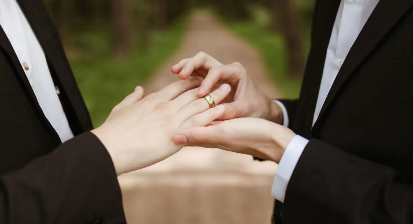 One person in a suit places a ring on anothers finger outdoors, symbolizing a wedding or engagement.