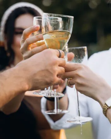 Four people clink wine glasses together outdoors, celebrating. Only their hands and glasses are visible in the close-up shot.