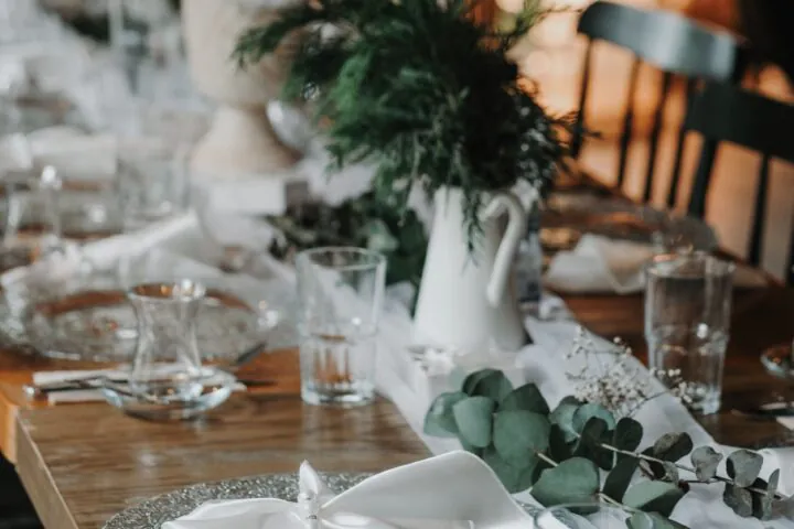 Elegant table setting with white napkins, clear glassware, and green foliage centerpieces on a wooden table in a softly lit room.