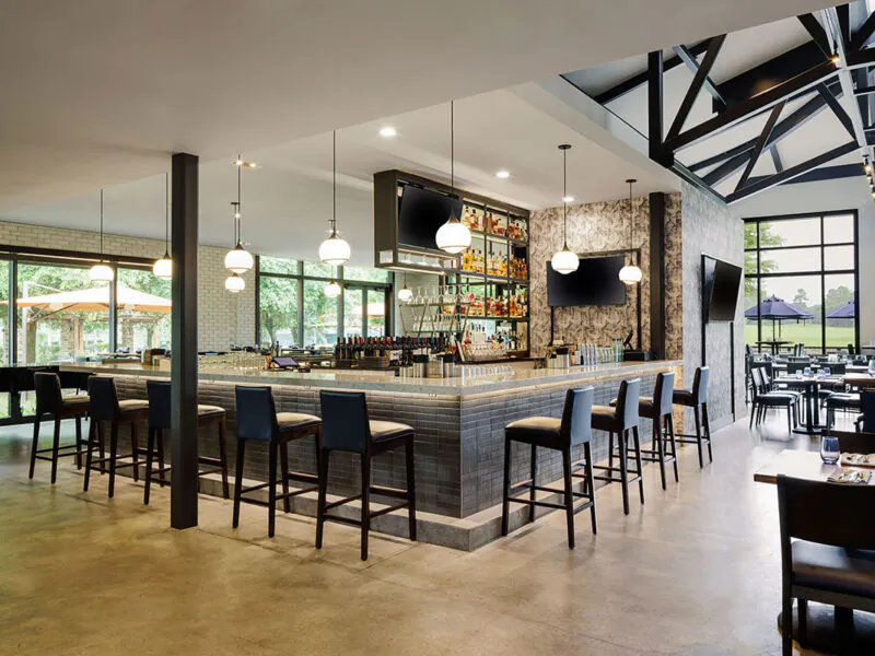 Modern restaurant interior with a central bar area, high chairs, hanging lights, large windows, and outdoor patio seating visible through the glass.