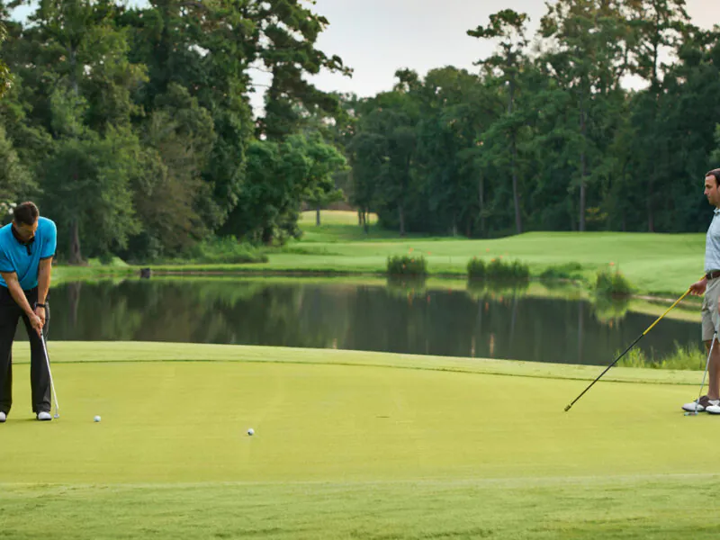 Two men on a golf green by a pond; one prepares to putt while the other holds a flagstick and watches.