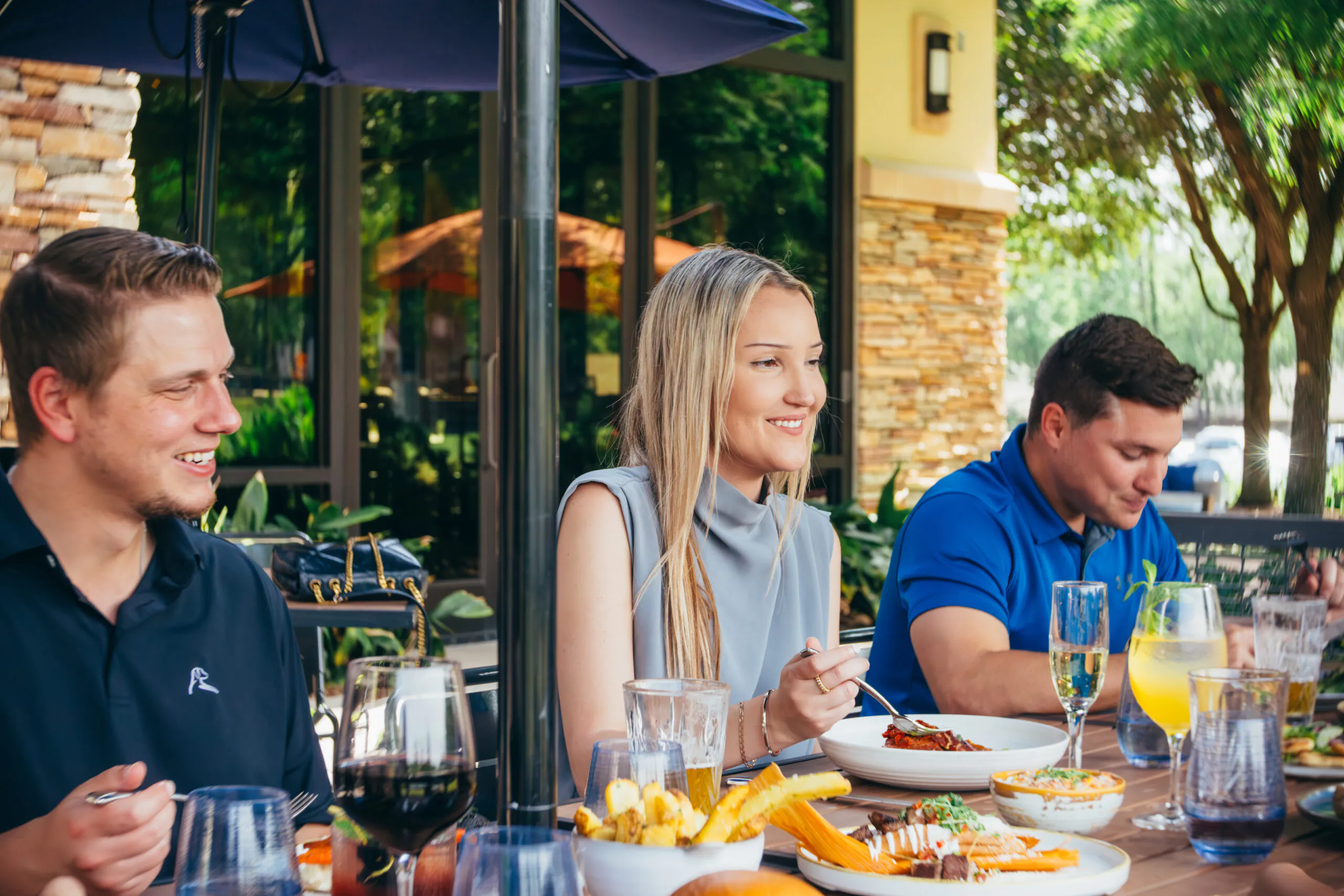 Three people sit at an outdoor restaurant table, eating and drinking, with food and drinks arranged in front of them on a sunny day.