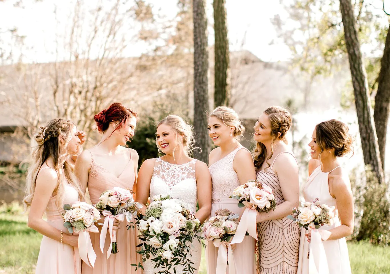A bride in a white dress stands outdoors with five bridesmaids in pale pink dresses, all holding bouquets and smiling at each other.