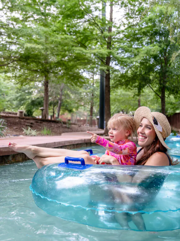 A woman and a child float in a blue tube on a lazy river, smiling and enjoying a sunny day surrounded by trees.