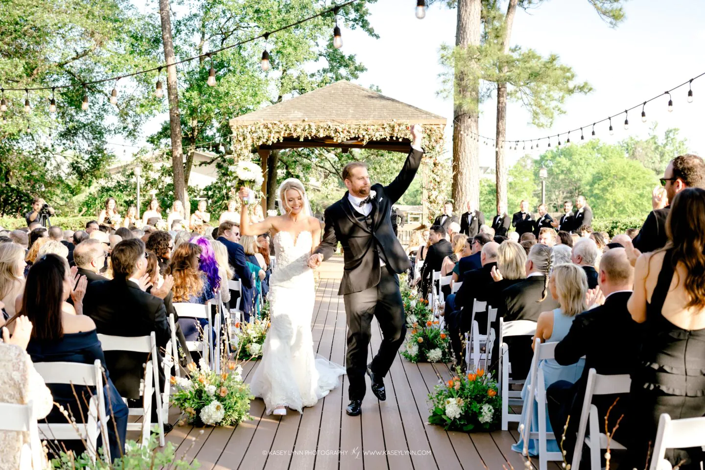 A bride and groom celebrate as they walk down the aisle outside, smiling and holding hands, surrounded by seated guests and floral decorations.