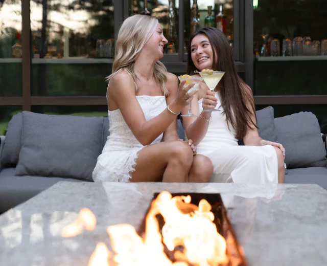 Two young women in white dresses sit on an outdoor sofa and toast with cocktails beside a fire pit.